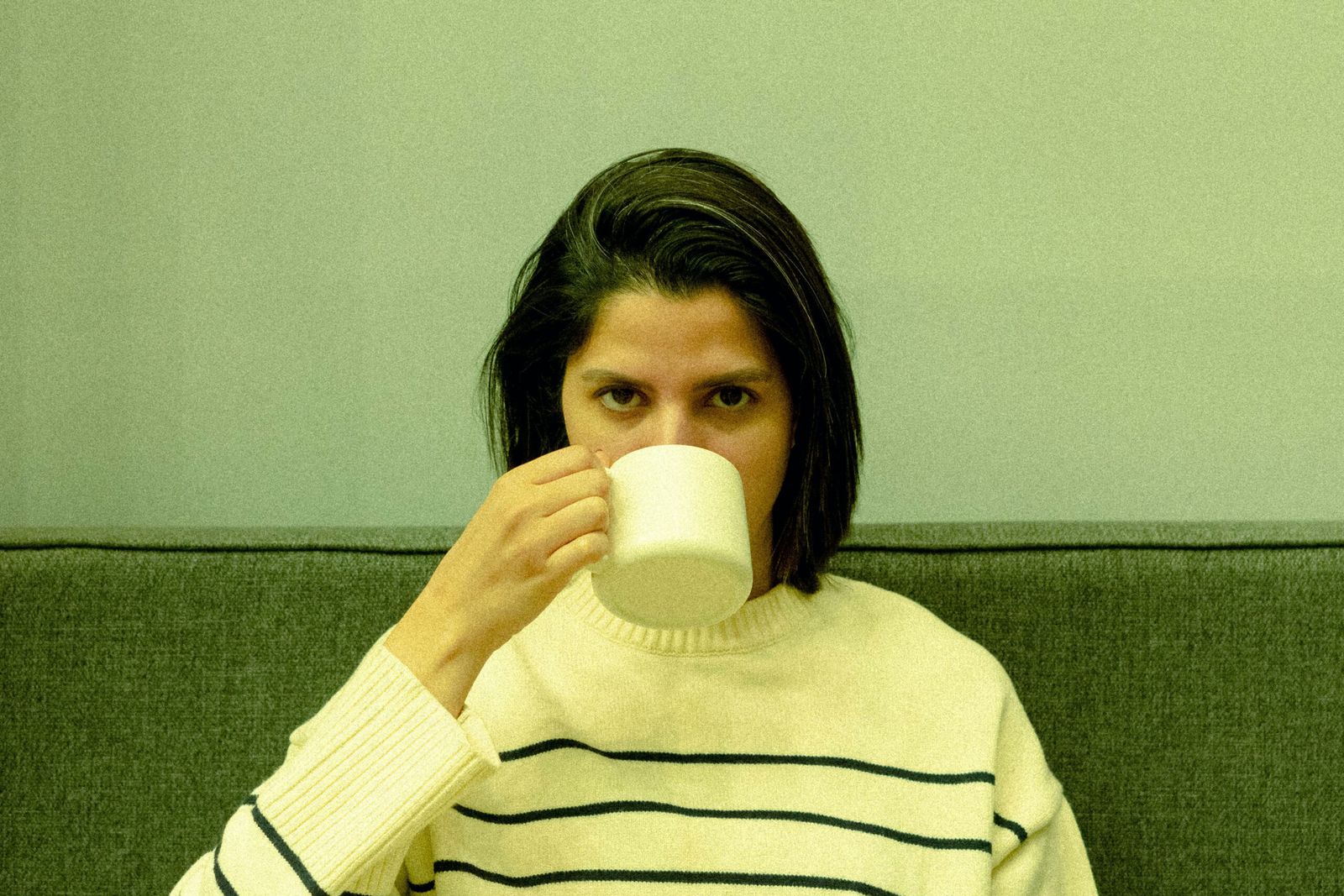 A young woman drinking from a white mug against a green wall